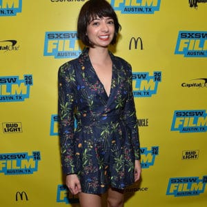 Kate Micucci wearing a blue wrap dress standing in front of a yellow background