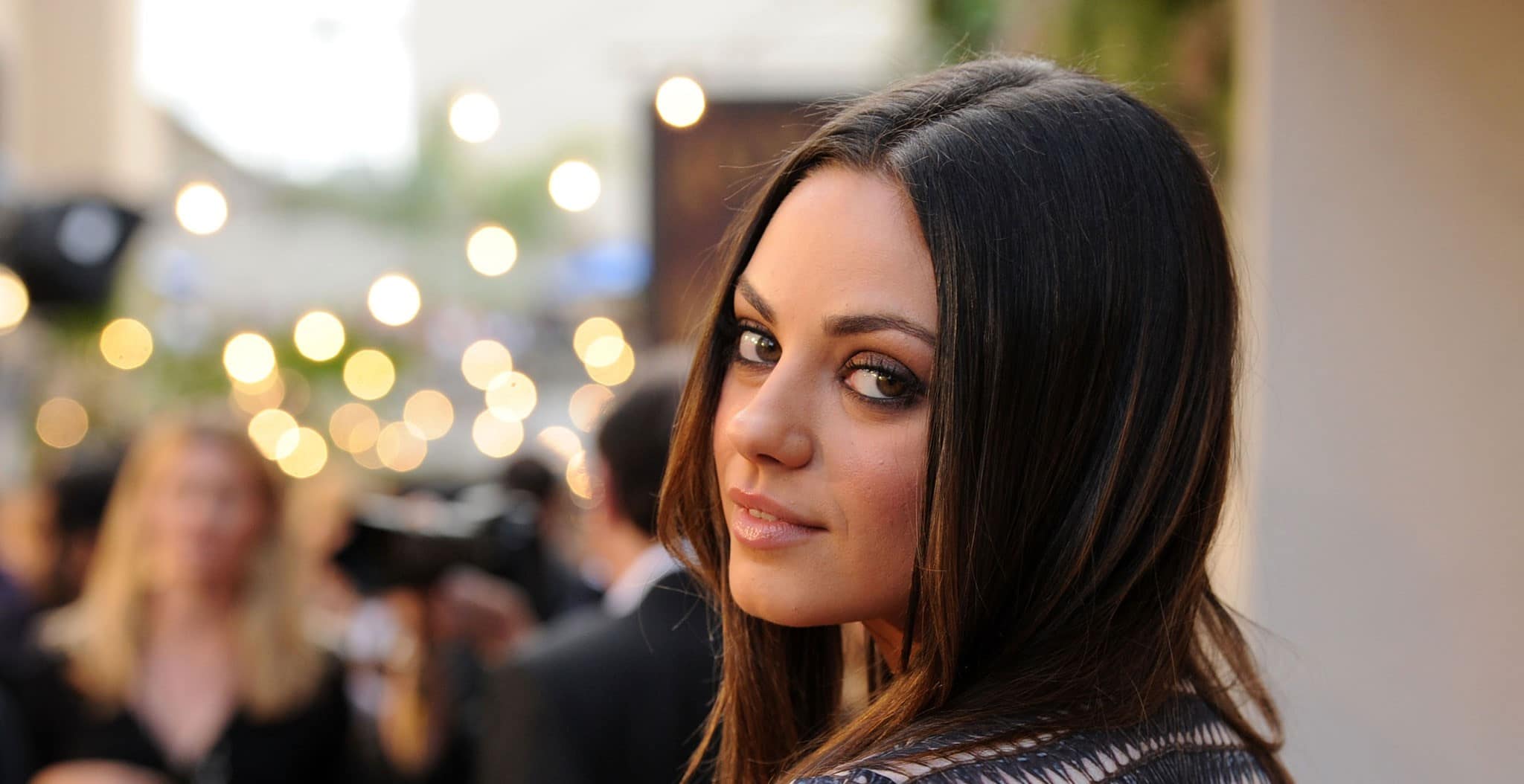 Mila Kunis looking back at the camera with lights in the background